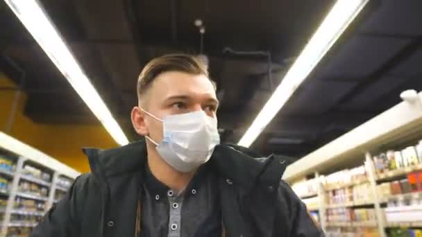 Porträt eines jungen Mannes mit medizinischer Gesichtsmaske, der im Supermarkt spazieren geht. Guy geht mit Schutzmaske vor dem Virus in den Laden. Einkaufen im Lebensmittelgeschäft während der Coronavirus-Pandemie. Gesundheitsbegriff. COVID-19 — Stockvideo