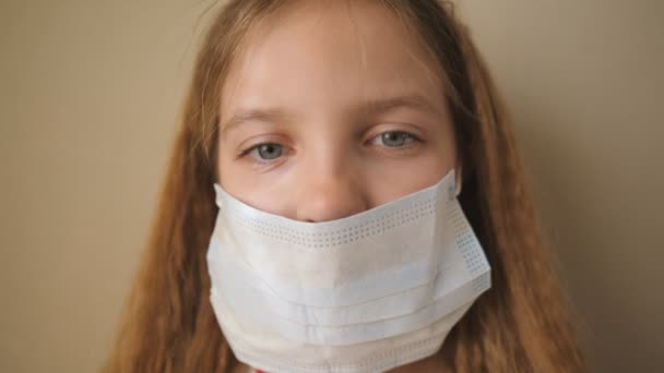 Portret van een klein meisje met medisch gezichtsmasker tijdens zelfisolatie. Triest vrouwelijk kind dragen beschermende masker van virus indoor. Concept van gezondheid en veiligheid van coronavirus en pandemie. Sluiten. — Stockvideo