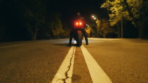 Motociclista en un casco y chaqueta de cuero está sentado en su motocicleta en la carretera de la noche y comenzar a montar. El hombre empieza a andar en moto por la noche. Concepto de libertad y aventura. Vista trasera — Vídeos de Stock