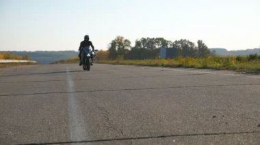 Genç adam boş otoyolda yavaşça spor motosikletine biniyor. Motosikletliler asfalt yolda düşük hızda gidiyorlar. Adam eğleniyor. Özgürlük ve macera kavramı. Düşük açı görünümü