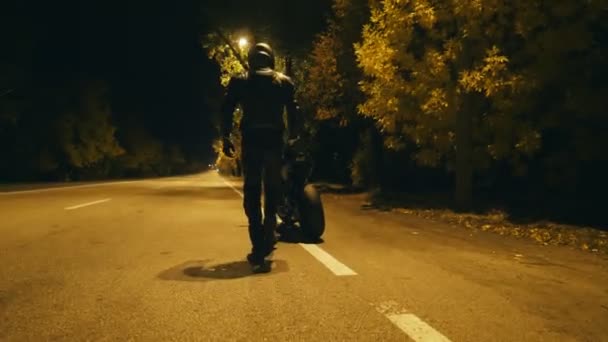 Motociclista em um capacete e jaqueta de couro que vem para sua motocicleta, senta-se nele e iniciando passeio na estrada noturna. O homem começa a andar de moto à noite. Conceito de liberdade e aventura. Vista traseira — Vídeo de Stock