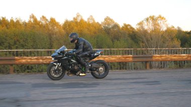 Kasklı motosikletçi, otoyoldaki eski paslı köprüde modern spor motosikletle hızlı gidiyor. Genç adam kır yolunda motosikletiyle yarışıyor. Bisikletli adam. Kıyametten sonraki kavram. Yan görünüm.