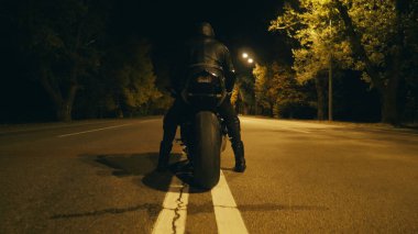 Kasklı ve deri ceketli bisikletçi gece otobanında motosikletinin üstünde ve sürmeye hazır. Adam akşam vakti motosiklete binecek. Özgürlük ve macera kavramı. Arka görünüm.