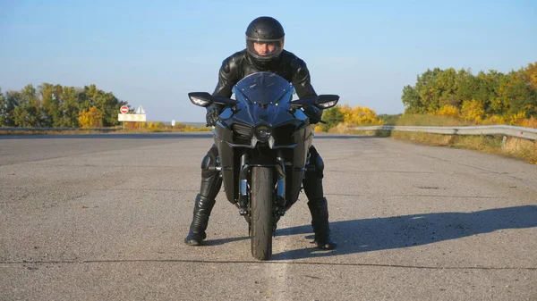 Front view of young biker in helmet and leather jacket is sitting on motorcycle at highway road and ready to ride. Man is going to ride at sport motorbike. Concept of freedom and adventure. Close up.