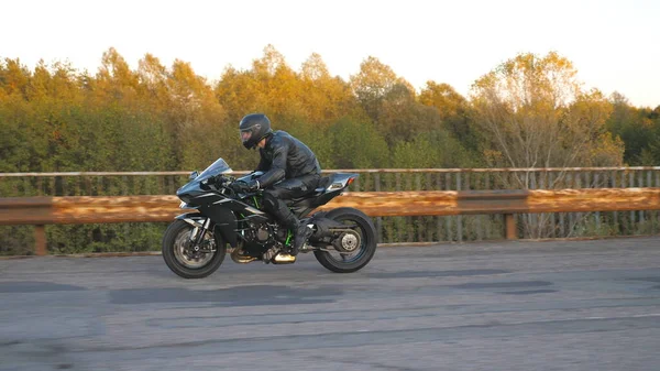 Kasklı motosikletçi, otoyoldaki eski paslı köprüde modern spor motosikletle hızlı gidiyor. Genç adam kır yolunda motosikletiyle yarışıyor. Bisikletli adam. Kıyametten sonraki kavram. Yan görünüm.