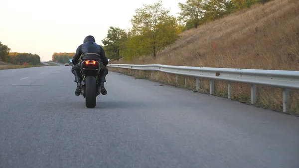 Sonbahar otobanında modern motosiklet kullanan tanınmayan bir adam. Motosikletli, kır yolunda motosikletiyle yarışıyor. Seyahat sırasında motosiklet sürücüsü. Özgürlük ve macera kavramı. Arka görünüm.