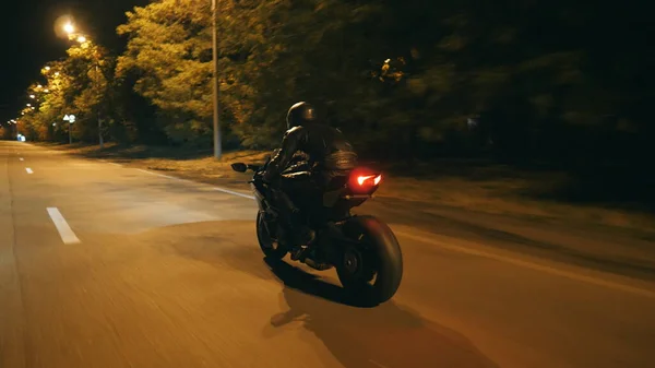 Motosikletli, akşam saatlerinde motosikletiyle yarışıyor. Şehrin gece caddesinde modern motosiklet süren bir adam. Yolculuk sırasında bisiklet süren adam. Özgürlük ve macera kavramı. Arka görünüm Kapat.