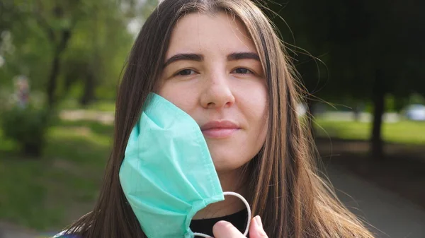 Mutlu genç bir kadının portresi tıbbi maskesini çıkarır ve gülümser. Kız dışarıdaki COVID 19 virüsünden koruyucu maskeyi çıkarıyor. Koronavirüs salgınından sağIık ve güvenlik kavramı. Kapat.