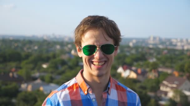 Portrét mladého šťastného muže sedícího na střeše vysoké budovy a hledícího do kamery s rozmazaným městským pozadím. Fešák si sundává sluneční brýle a směje se. Zavřít Zpomalený pohyb — Stock video