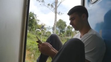 Erkek turist hareket halindeki trenin açık kapısında oturur ve akıllı telefon kullanır. Genç adam yolcu trenine biniyor ve cep telefonundaki fotoğraflara bakıyor. Yaz seyahati ya da turizm kavramı. Yavaş çekim