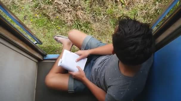 Top view of young man sits near entrance of moving train and looks out at nature. Male tourist rides on passenger railway transport and holds in hands interesting book. Concept of vacation — Stock Video