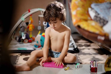 Kıvırcık saçlı, şortlu çocuk, kıyafetlerini kirletmemek için odasında sümükle oynuyor..