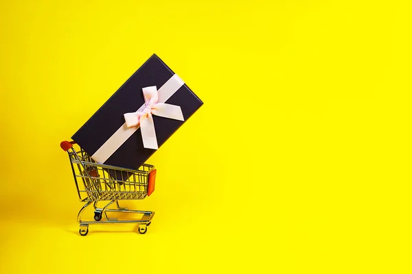 Regalo de vacaciones, concepto de Viernes Negro con caja de regalo y lazo rosa en un carrito de compras de metal, canasta sobre fondo de color amarillo . — Foto de Stock