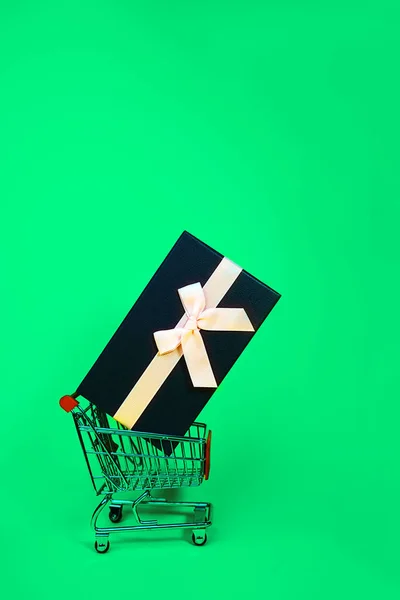 Regalo navideño, concepto Black Green Friday con caja de regalo y lazo rosa en un carrito de compras de metal, canasta sobre fondo de color verde . — Foto de Stock