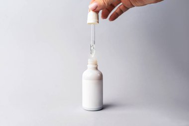 Elinde bir damla serum ya da hyaluronik asit ya da kolajen nemlendirici ve gri arka planda beyaz şişe olan bir kadın eli.