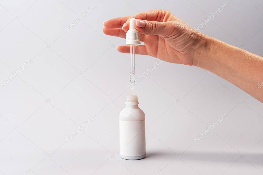 Hand of woman with pipette with drop of serum or hyaluronic acid or collagen moisturizer and white bottle on gray background.