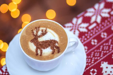  Beyaz bardakta kapuçino, içinde Noel geyiği latte sanatı var.