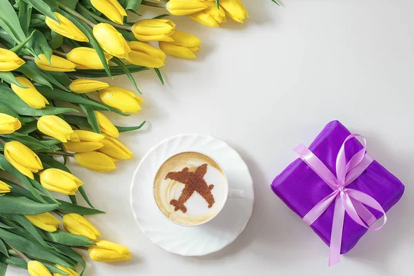cup of cappuccino coffee with a pattern of airplane made of cinnamon on milk foam, yellow tulips and wrapped gift box