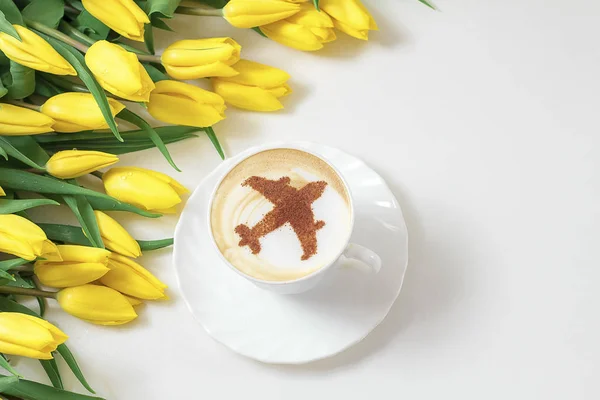cup of cappuccino coffee with a pattern of airplane made of cinnamon on milk foam, yellow tulips