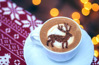  Beyaz bardakta kapuçino, içinde Noel geyiği latte sanatı var.