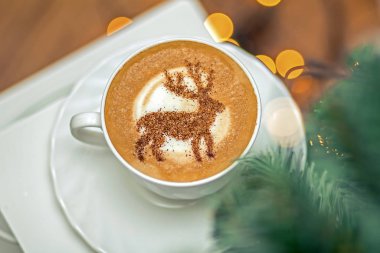  Beyaz bardakta kapuçino, içinde Noel geyiği latte sanatı var.
