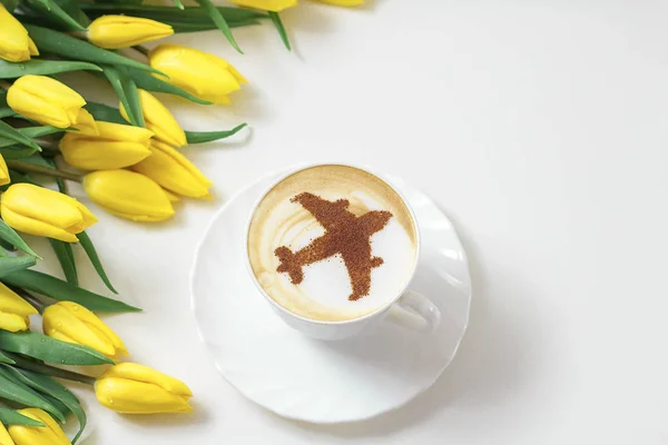 cup of cappuccino coffee with a pattern of airplane made of cinnamon on milk foam, yellow tulips