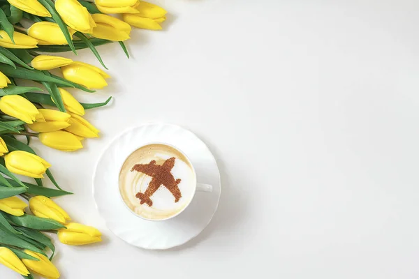 cup of cappuccino coffee with a pattern of airplane made of cinnamon on milk foam, yellow tulips