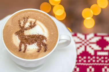  Beyaz bardakta kapuçino, içinde Noel geyiği latte sanatı var.