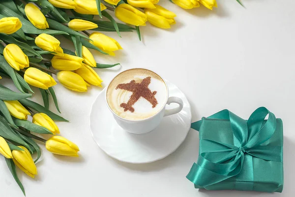 cup of cappuccino coffee with a pattern of airplane made of cinnamon on milk foam, yellow tulips and wrapped gift box