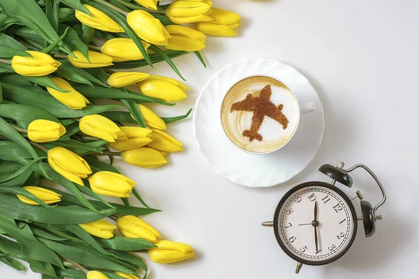 cup of cappuccino coffee with a pattern of airplane made of cinnamon on milk foam, yellow tulips and vintage alarm clock