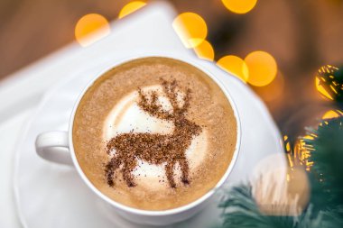  Beyaz bardakta kapuçino, içinde Noel geyiği latte sanatı var.