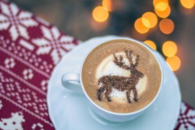  Beyaz bardakta kapuçino, içinde Noel geyiği latte sanatı var.