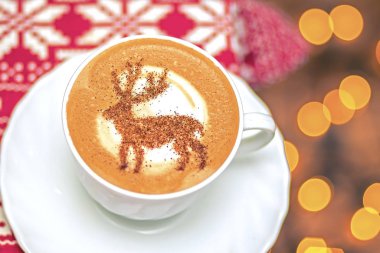  Beyaz bardakta kapuçino, içinde Noel geyiği latte sanatı var.