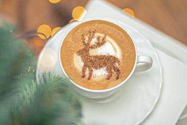  Beyaz bardakta kapuçino, içinde Noel geyiği latte sanatı var.