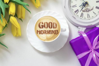 cup of cappuccino coffee with a pattern of GOOD MORNING made of cinnamon on milk foam, yellow tulips, alarm clock and gift box