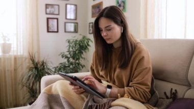 Kahverengi kazaklı, elinde akıllı bir saat ve kablosuz kulaklıkla genç ve güzel bir kız tabletteki postaları tarıyor.