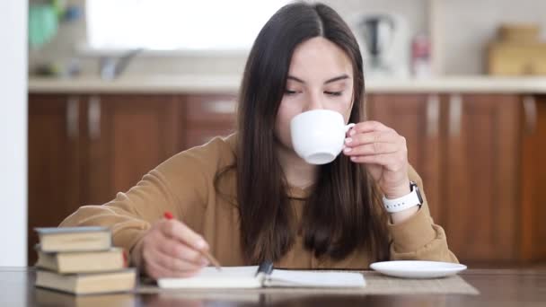 茶色いセーターの少女がノートにメモを書き 台所のテーブルに座ってお茶を飲む — ストック動画