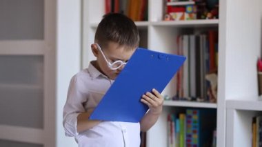 Beyaz gözlüklü küçük bir çocuk doktoru oynuyor ve not defterine notlar yazıyor.