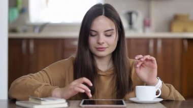 Kahverengi kazaklı bir kız tablet kullanıyor ve mutfak masasında otururken çay içiyor.