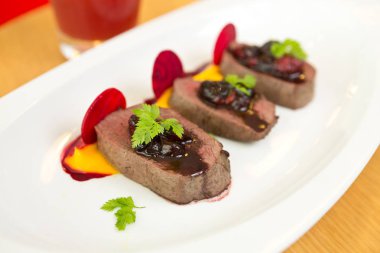 Organik Geyik eti beyaz bir tabak üzerinde taze ahududu ahududu sosu, pancar dilimleri ve kabak püresi ile servis.