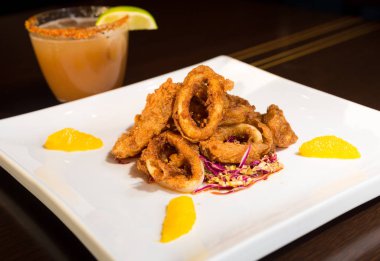 Güzel altın gevrek derin kızarmış kalamar halkaları, turuncu kesimleri ve arka planda yak²n bir kokteyl ile beyaz bir tabakta servis.