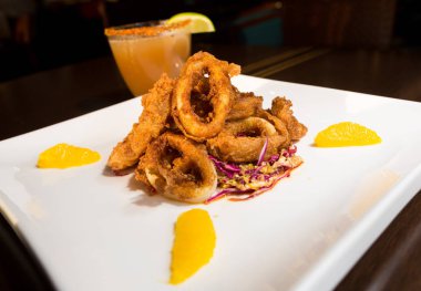 Güzel altın gevrek derin kızarmış kalamar halkaları, turuncu kesimleri ve arka planda yak²n bir kokteyl ile beyaz bir tabakta servis.