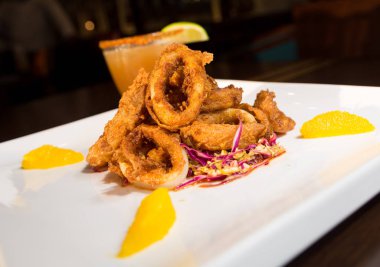 Güzel altın gevrek derin kızarmış kalamar halkaları, turuncu kesimleri ve arka planda yak²n bir kokteyl ile beyaz bir tabakta servis.