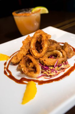 Güzel altın gevrek derin kızarmış kalamar halkaları, turuncu kesimleri ve arka planda yak²n bir kokteyl ile beyaz bir tabakta servis.