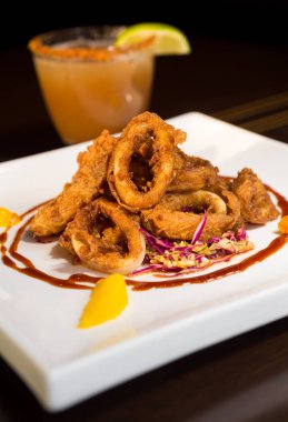 Güzel altın gevrek derin kızarmış kalamar halkaları, turuncu kesimleri ve arka planda yak²n bir kokteyl ile beyaz bir tabakta servis.