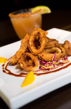 Güzel altın gevrek derin kızarmış kalamar halkaları, turuncu kesimleri ve arka planda yak²n bir kokteyl ile beyaz bir tabakta servis.