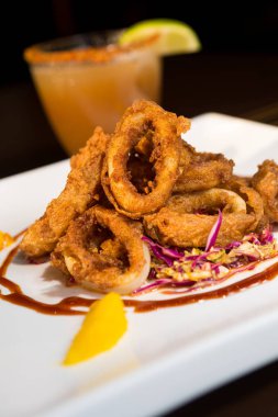 Güzel altın gevrek derin kızarmış kalamar halkaları, turuncu kesimleri ve arka planda yak²n bir kokteyl ile beyaz bir tabakta servis.