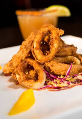 Güzel altın gevrek derin kızarmış kalamar halkaları, turuncu kesimleri ve arka planda yak²n bir kokteyl ile beyaz bir tabakta servis.