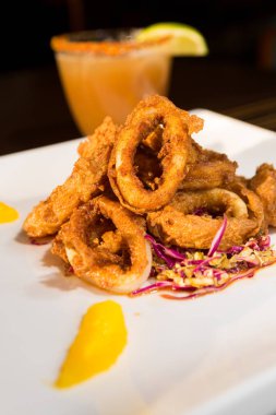 Güzel altın gevrek derin kızarmış kalamar halkaları, turuncu kesimleri ve arka planda yak²n bir kokteyl ile beyaz bir tabakta servis.
