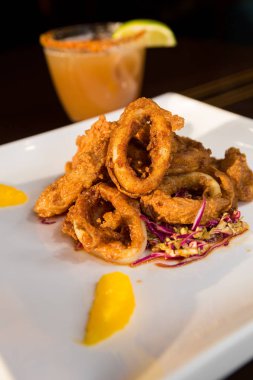 Güzel altın gevrek derin kızarmış kalamar halkaları, turuncu kesimleri ve arka planda yak²n bir kokteyl ile beyaz bir tabakta servis.
